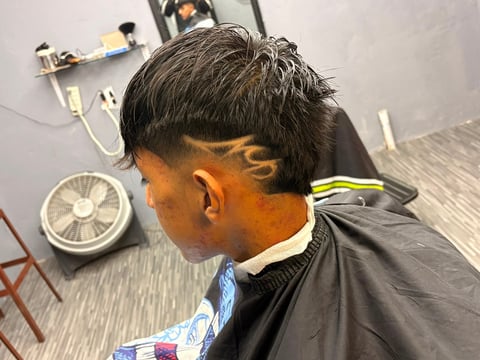 Persona mostrando un corte de barbería con diseño de llamas en la parte posterior y nuca, usando capa negra, fotografiada de perfil