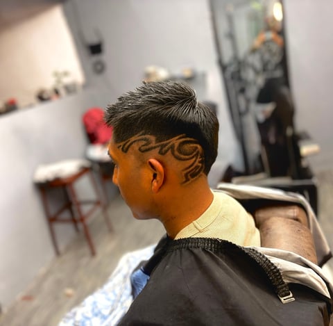 Hombre con tatuaje decorativo en la parte posterior de la cabeza y cuello en una barbería, usando capa negra