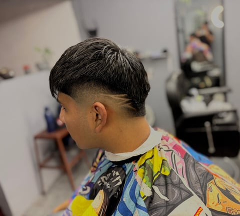 Joven con cabello oscuro corto y laterales degradados sentado en una silla de barbería, usando camisa colorida